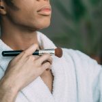 A man in a bathrobe applies foundation with a brush indoors.